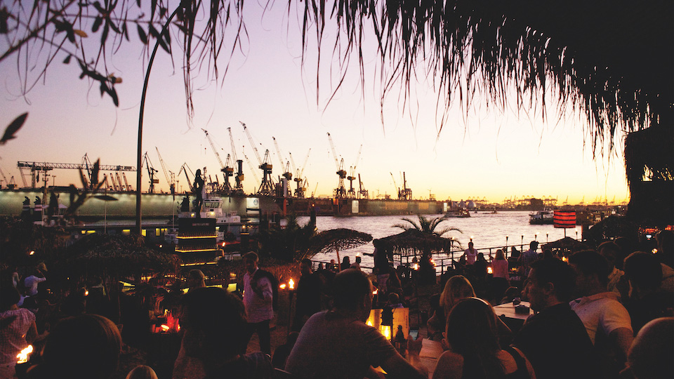 Menschen sitzen bei Sonnenuntergang in einem Hamburger Beachclub vor der Kulisse des Hafens