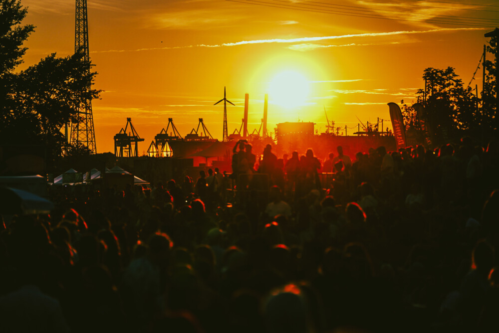 unkenntliche Menschenmasse zieht vor einer Hafen-Kulisse bei Sonnenuntergang über ein Festival Gelände 