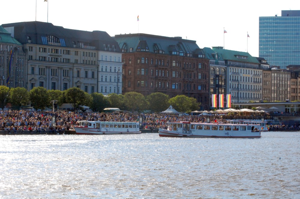 Blick auf den Jungfernstieg zur Hamburger Pride Week während des CSD-Straßenfest: viele Menschen säumen die Wasserfront, zwei Fähren im Vordergrund, Regenbogenflaggen im Hintergrund