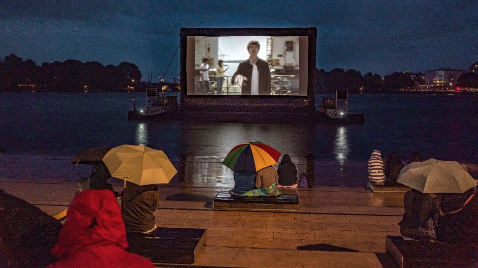 Eine Kinoleinwand ist auf der Binnenalster in Hamburg befestigt. Das Publikum sitzt unter Regenschirmen auf Treppen davor