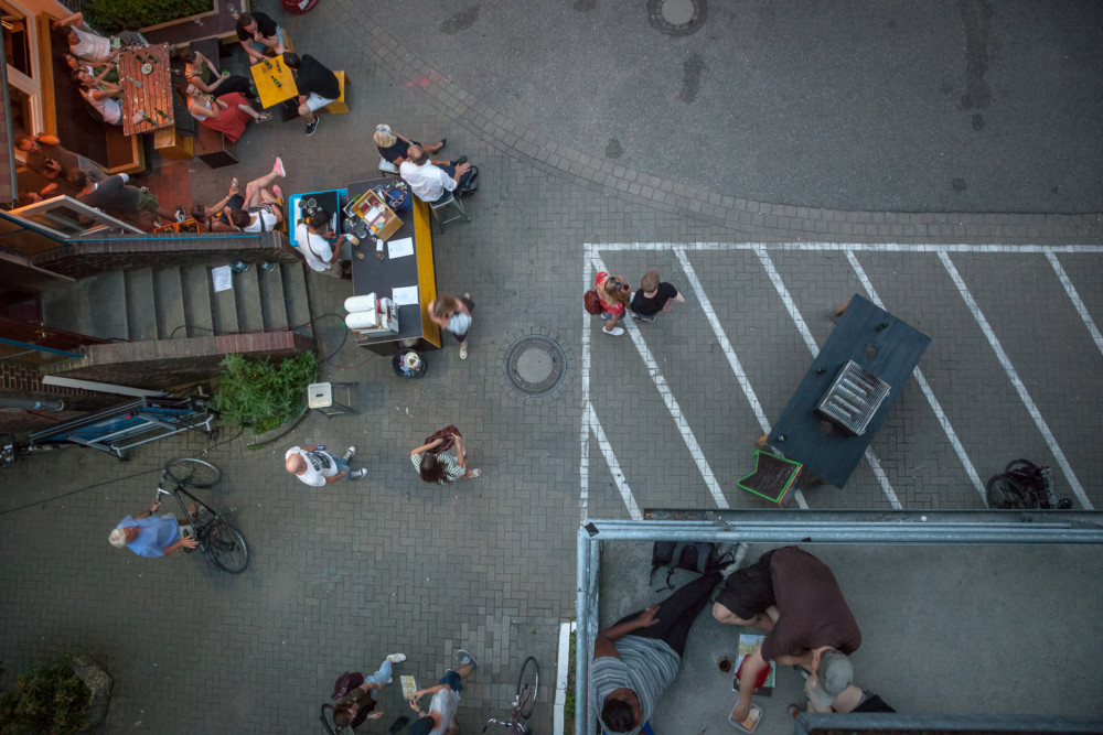 Menschen sitzen auf der Straße und auf einem Balkon, aus der Vogelperspektive.