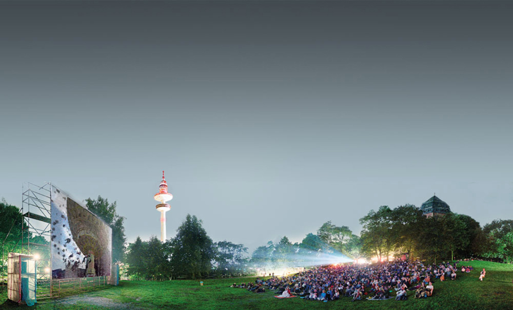 Panoramaaufnahme eines Open-Air-Kinos in Hamburg im Schanzenpark inklusive Leinwand, Publikum, Fernsehtum und Wasserturm