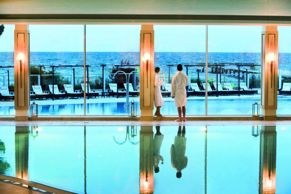 Zwei Menschen in weißen Bademäntel stehen an hohen Panorama-Fenstern zwischen Innen- und Außenpool eines Hotels mit Meerblick