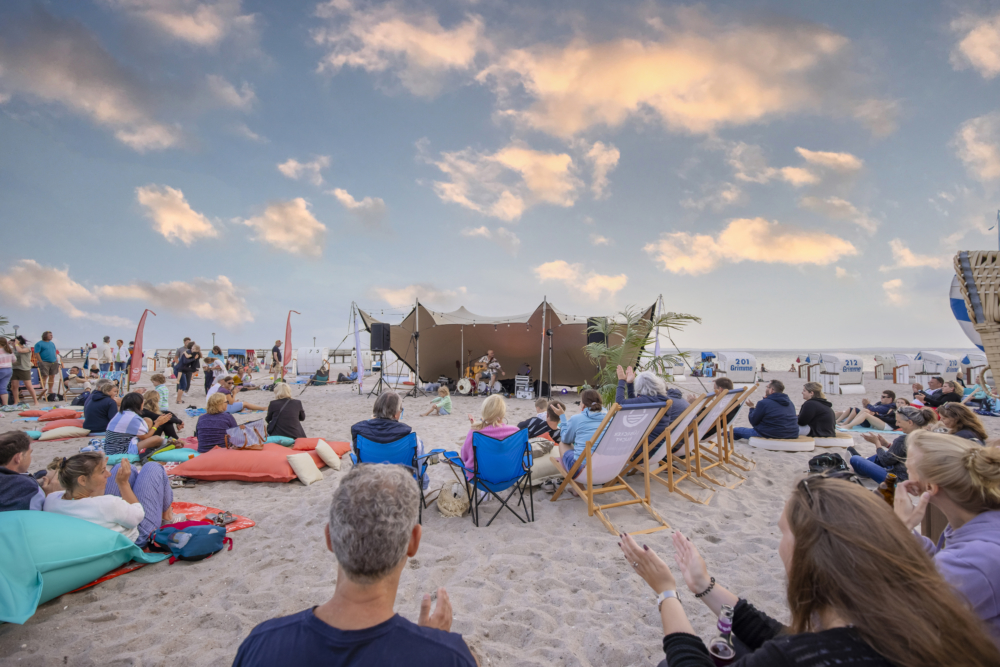 Konzert am Strand von Pelzerhaken: Die Zuschauenden sitzen auf Liegestühlen und Sitzkissen im Sand  