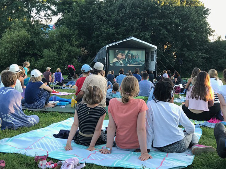 Kinder sitzen auf Decken vor einer Open-Air-Kinoleinwand in einem Park im Sommer