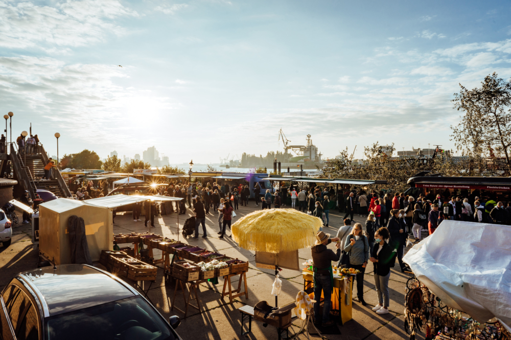 Stände vom Hamburger Fischmarkt im Sonnenschein. Im Hintergrund sieht man Hafenkrähne und die Elbphilharmonie. 