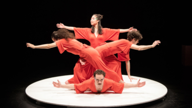 Vier Tänzer und Tänzerinnen in roten Anzügen formatieren sich auf einem weißen Kreis vor schwarzem Hintergrund.