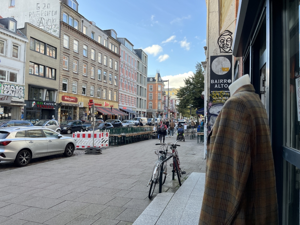 Im Vordergrund eine kopflose Schaufensterpuppe mit einem Sakko, im Hintergrund das Treiben des Schulterblatts, eine beleibte Einkaugstraße in Hamburg 