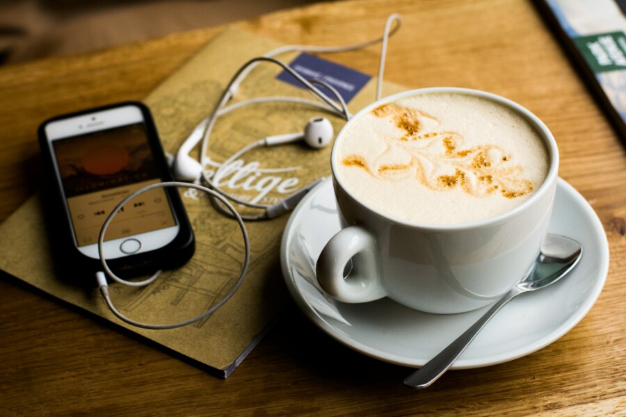 Tisch mit Cappuccino-Tasse daneben liegt ein Smartphone mit Kopfhörern