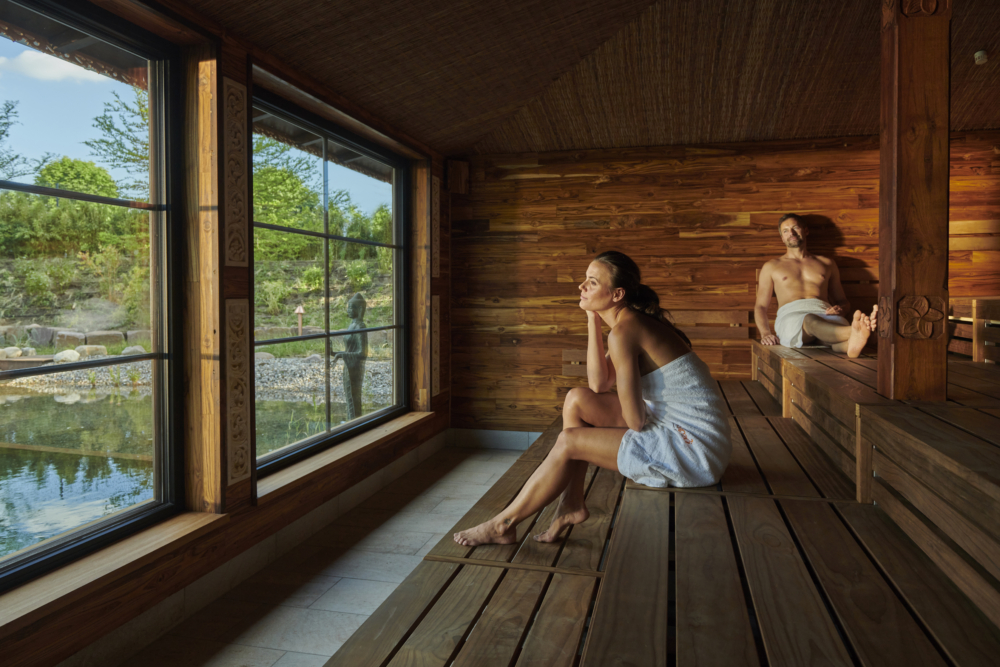 Zwei Menschen sitzen in einer Sauna von vabali spa und blicken aus den Panoramafenstern auf einen Teich