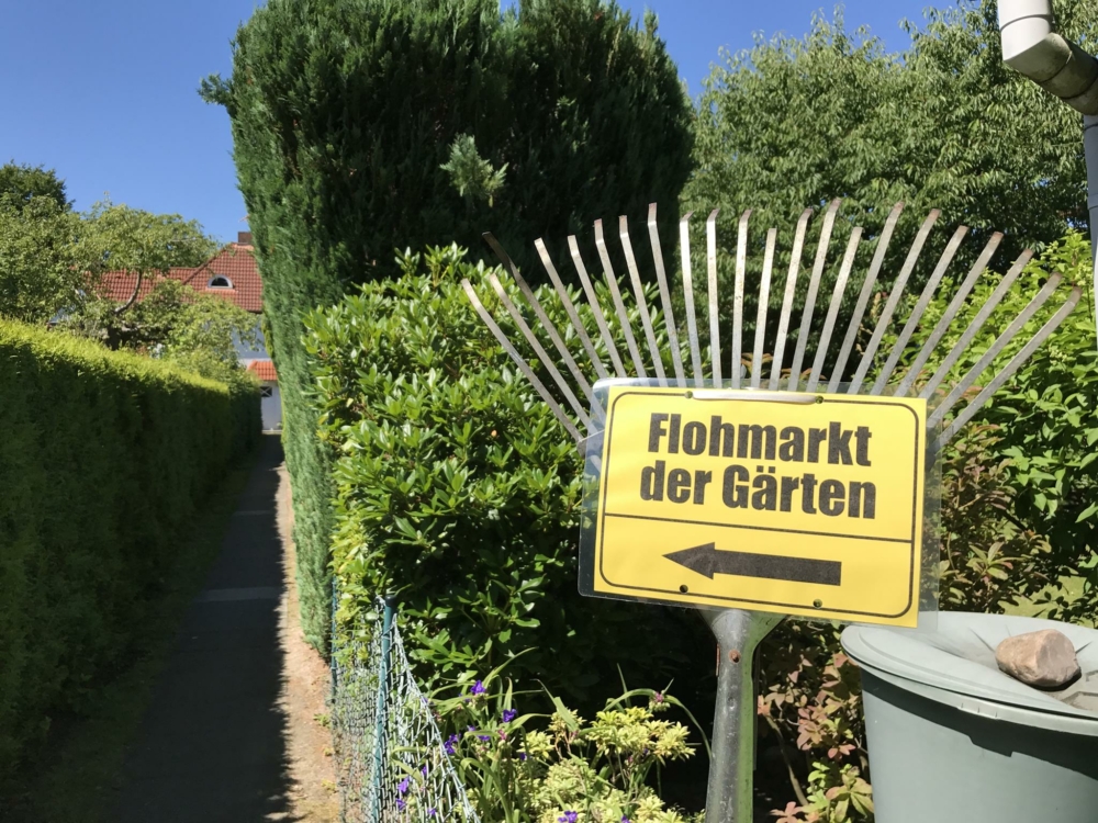 Zu sehen ist: Ein gelbes Schild auf dem mit schwarzer Schrift Flohmarkt der Gärten steht. Darunter befindet sich ein schwarzer Pfeil. Er soll darauf hinweisen, wo sich der Flohmarkt befindet.