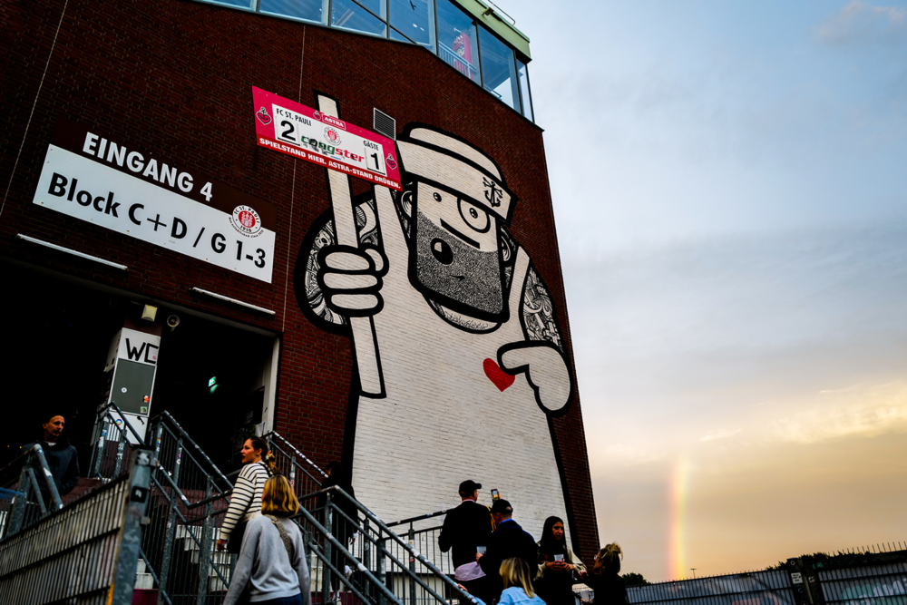 Eine Hausfassade des Stadions, auf der ein Graffiti zu sehen ist. Das Graffiti zeigt einen Seemann, der ein FC St. Pauli Ticket in den Händen hält und ein rotes Herz auf seine Brust gemalt hat.