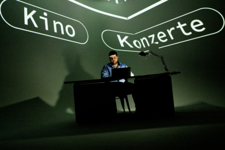 Ersin sitzt auf der Bühne und bedient seinen Laptop. Im Hintergrund stehen die Worte "Kino" und "Konzert" an die Wand geworfen.