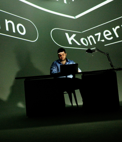 Ersin sitzt auf der Bühne und bedient seinen Laptop. Im Hintergrund stehen die Worte "Kino" und "Konzert" an die Wand geworfen.