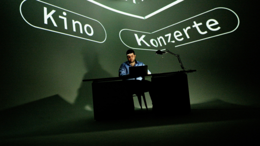Ersin sitzt auf der Bühne und bedient seinen Laptop. Im Hintergrund stehen die Worte "Kino" und "Konzert" an die Wand geworfen.