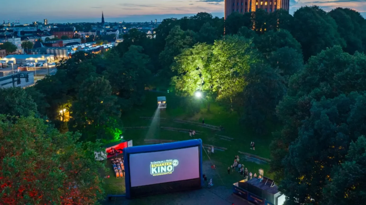Freiluftkino mit aufblasbarer Leinwand und Zuschauern im Park bei Abenddämmerung, im Hintergrund ein beleuchteter Wasserturm