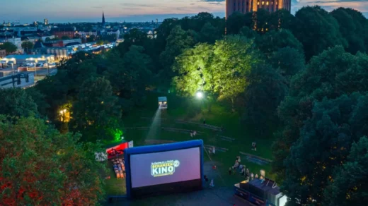 Freiluftkino mit aufblasbarer Leinwand und Zuschauern im Park bei Abenddämmerung, im Hintergrund ein beleuchteter Wasserturm