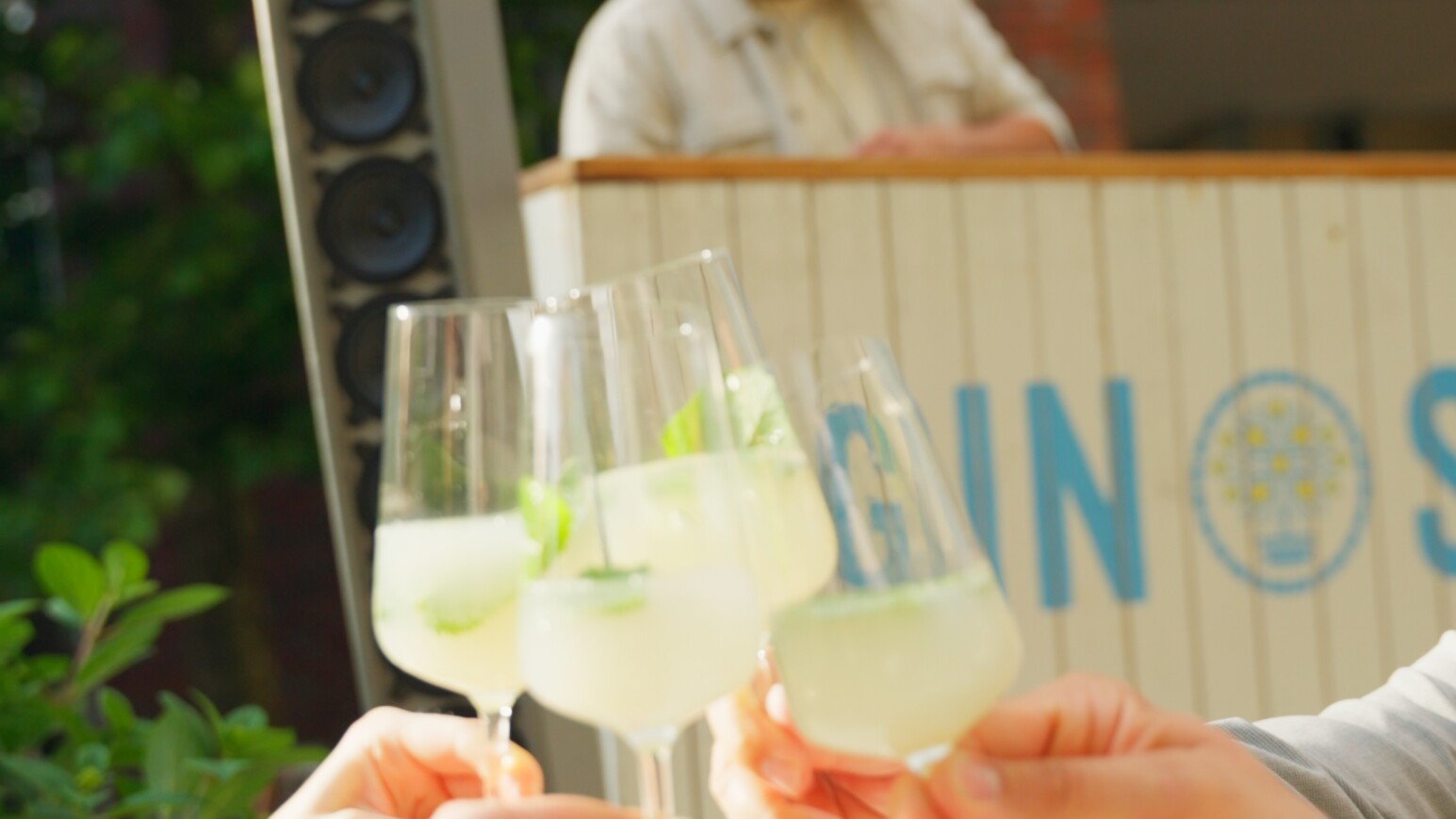 Vier Hände stoßen mit Cocktails an, im Hintergrund spielt ein DJ Musik.