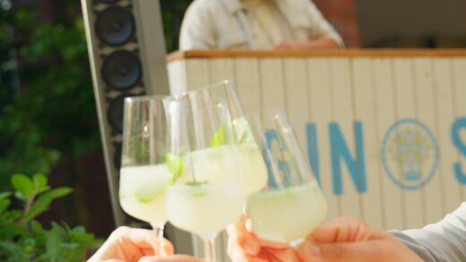 Vier Hände stoßen mit Cocktails an, im Hintergrund spielt ein DJ Musik.
