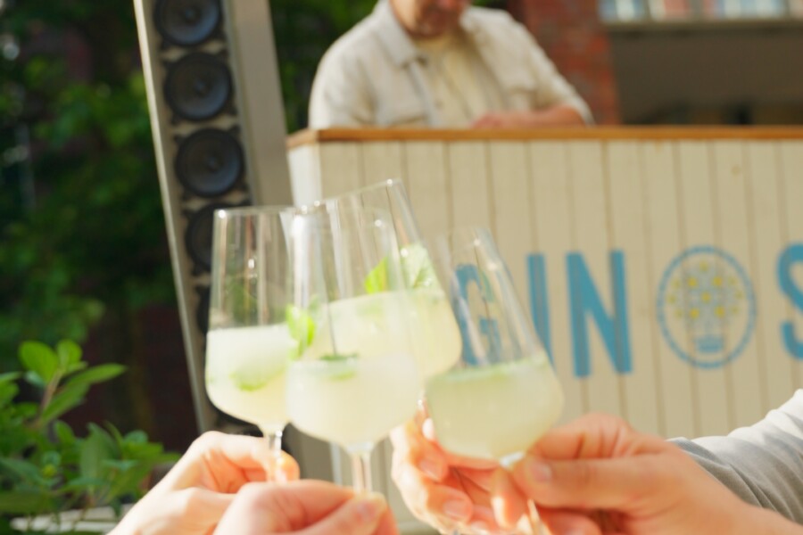 Vier Hände stoßen mit Cocktails an, im Hintergrund spielt ein DJ Musik.