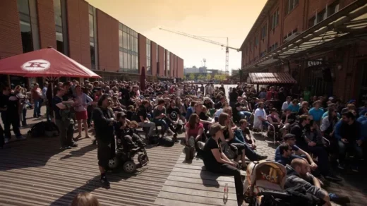 Große Menschenmenge sitzt und steht auf Holzplanken zwischen modernen Gebäuden beim Konzert am Lattenplatz bei Sonnenuntergang