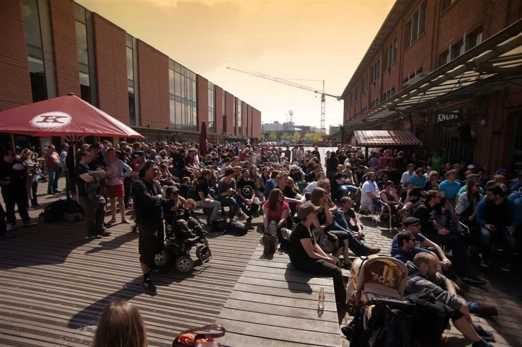 Große Menschenmenge sitzt und steht auf Holzplanken zwischen modernen Gebäuden beim Konzert am Lattenplatz bei Sonnenuntergang