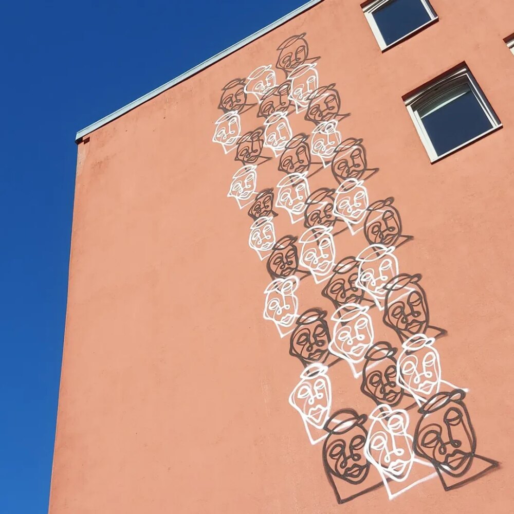 Lachsfarbene Hauswand, von unten nach oben aufgenommen. Rechts zwei Fenster, links daneben verlaufen Reihen markanter Gesichter übereinander, die sich von oben fast bis nach unten ziehen.