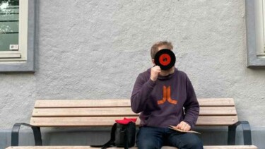 DJ Nico Thien sitzt auf einer Holzbank vor einer grauen Wand, trägt einen dunkelblauen Pullover mit orangefarbenem Logo und schwarze Schuhe, neben ihm eine schwarze Tasche mit rotem Innenfutter