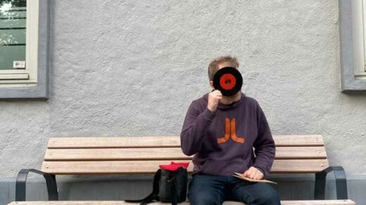 DJ Nico Thien sitzt auf einer Holzbank vor einer grauen Wand, trägt einen dunkelblauen Pullover mit orangefarbenem Logo und schwarze Schuhe, neben ihm eine schwarze Tasche mit rotem Innenfutter