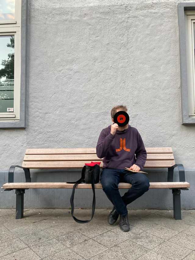 DJ Nico Thien sitzt auf einer Holzbank vor einer grauen Wand, trägt einen dunkelblauen Pullover mit orangefarbenem Logo und schwarze Schuhe, neben ihm eine schwarze Tasche mit rotem Innenfutter