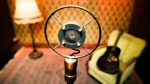 Nahaufnahme eines Vintage-Rundfunkmikrofons mit Ringhalterung vor einem Sessel mit Gitarre und einer Stehlampe