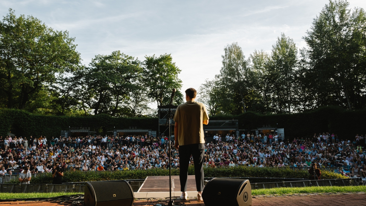 Poetry Slam Teilnehmer steht auf Bühne vor großem Publikum im Freien, umgeben von Bäumen