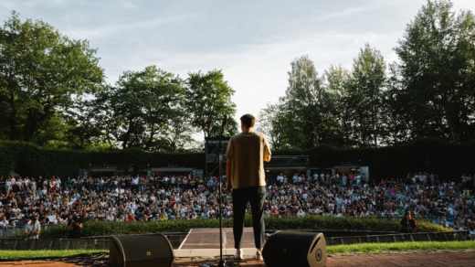 Poetry Slam Teilnehmer steht auf Bühne vor großem Publikum im Freien, umgeben von Bäumen