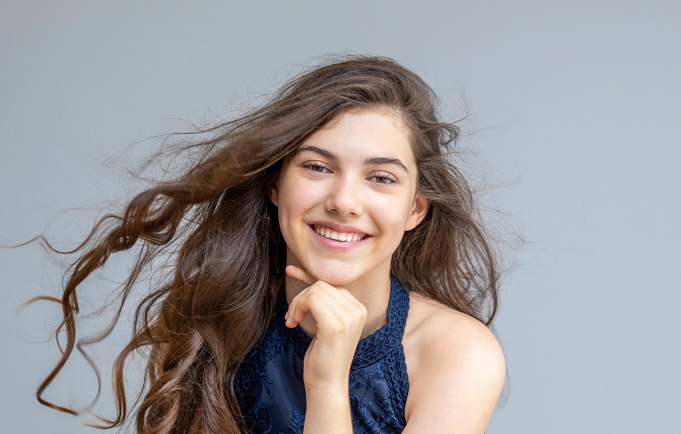 İlyun Bürkev mit langen braunen Haaren, sitzend, trägt ein ärmelloses blaues Spitzenkleid, Hand an Kinn