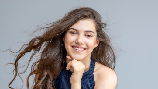 İlyun Bürkev mit langen braunen Haaren, sitzend, trägt ein ärmelloses blaues Spitzenkleid, Hand an Kinn