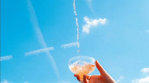 Hand hält ein Sektglas, in das aus einer Flasche rosafarbener Sekt eingeschenkt wird, blauer Himmel im Hintergrund