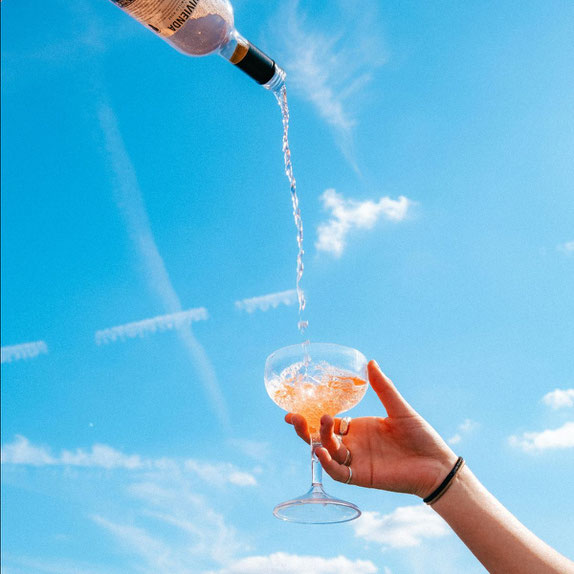 Hand hält ein Sektglas, in das aus einer Flasche rosafarbener Sekt eingeschenkt wird, blauer Himmel im Hintergrund