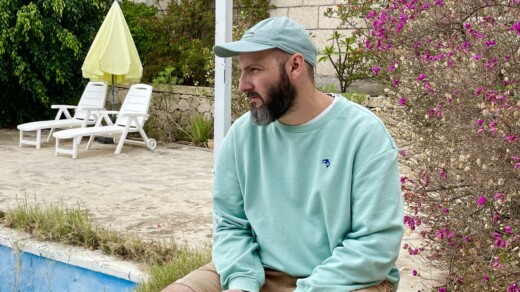 Ein Mann auf einem Stuhl - im Hintergrund ein Busch und ein Pool an dem zwei Liegen stehen