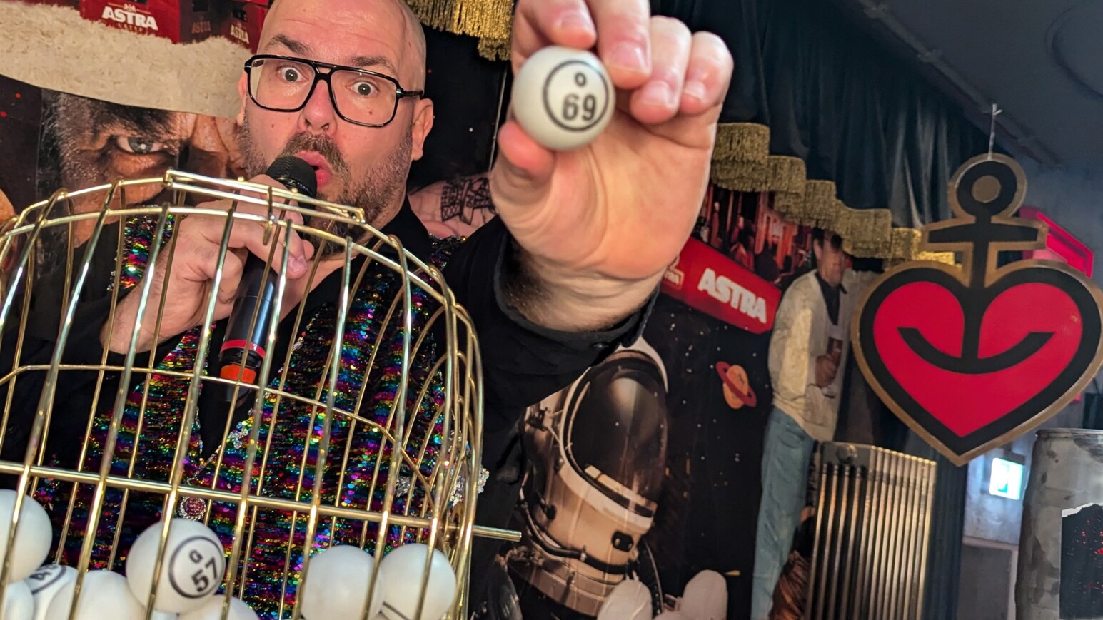 Hand hält eine Bingo-Kugel mit der Nummer 69 vor einem goldfarbenen Bingo-Korb mit weiteren Kugeln, Hintergrund mit Astra-Logo und einem roten Herz mit Anker-Symbol