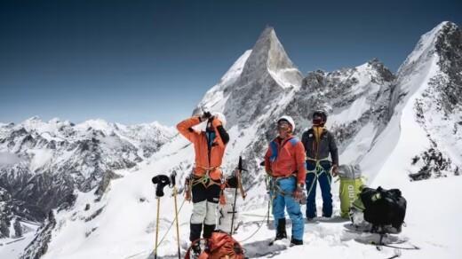 Drei Bergsteiger in bunter Ausrüstung stehen auf schneebedecktem Gipfel mit markantem spitzen Berg im Hintergrund