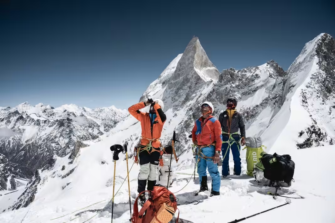 Drei Bergsteiger in bunter Ausrüstung stehen auf schneebedecktem Gipfel mit markantem spitzen Berg im Hintergrund