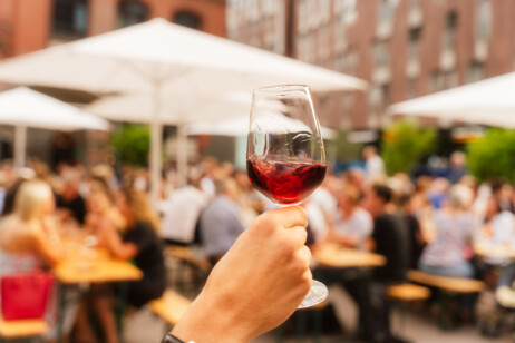 Vordergrund - Ein Wein der gehalten wird und im Hintergrund - Personen auf Bierbänken