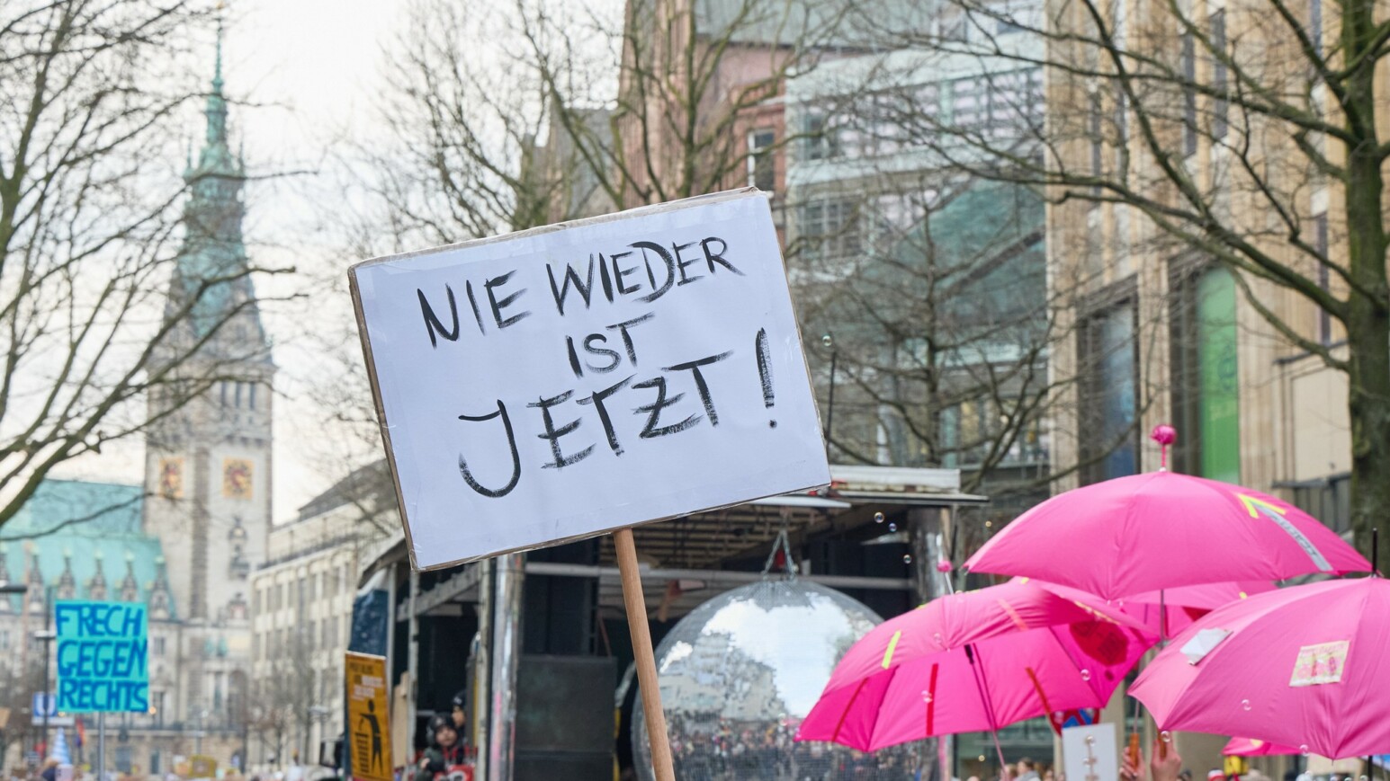 Protestplakat mit der Aufschrift 'NIE WIEDER IST JETZT!' vor einer Gruppe von Menschen mit pinken Regenschirmen in einer Stadt