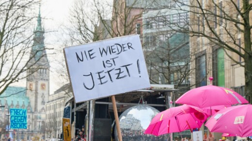 Protestplakat mit der Aufschrift 'NIE WIEDER IST JETZT!' vor einer Gruppe von Menschen mit pinken Regenschirmen in einer Stadt