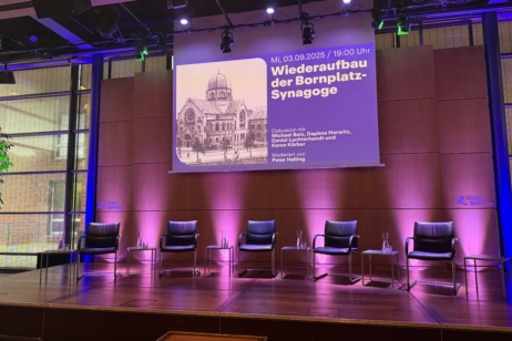 Bühne mit 5 leeren Stühlen für Podiumsdiskussion vorbereitet, darüber eine Leinwand mit dem Thema des Abends abgebildet