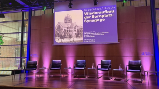 Bühne mit 5 leeren Stühlen für Podiumsdiskussion vorbereitet, darüber eine Leinwand mit dem Thema des Abends abgebildet