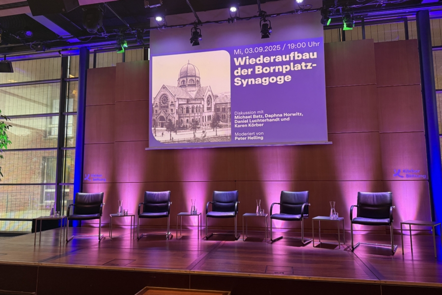 Bühne mit 5 leeren Stühlen für Podiumsdiskussion vorbereitet, darüber eine Leinwand mit dem Thema des Abends abgebildet