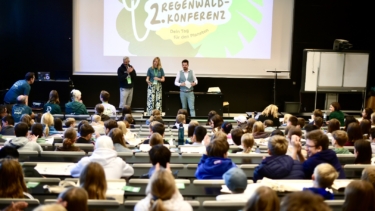 Vorlesesaal mit drei Personen vorne auf der Bühne, hinter ihnen eine Leinwand mit einem Bild. In den Sitzreihen sitzen viele Kinder mit dem Rücken zur Kamera.