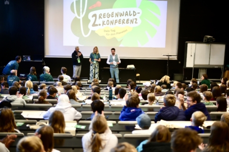 Vorlesesaal mit drei Personen vorne auf der Bühne, hinter ihnen eine Leinwand mit einem Bild. In den Sitzreihen sitzen viele Kinder mit dem Rücken zur Kamera.