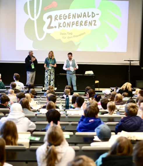 Vorlesesaal mit drei Personen vorne auf der Bühne, hinter ihnen eine Leinwand mit einem Bild. In den Sitzreihen sitzen viele Kinder mit dem Rücken zur Kamera.